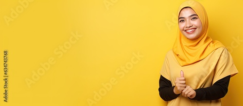 A happy Asian Muslim woman pointing to the left with copy space standing on a yellow background © HN Works