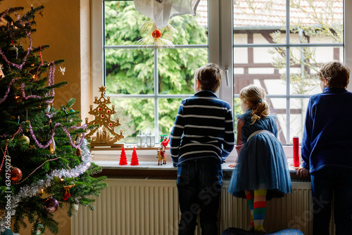 Three cute children sitting by window on Christmas eve. Two school kid boys and toddler girl, siblings looking outdoor and dreaming. Family happiness on traditional holiday