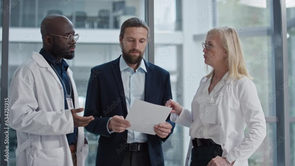 Medium shot of young entrepreneur discussing strategy of clinic growth and showing something in document to doctors Medium shot of young entrepreneur discussing strategy of clinic growth and showing something in document to doctors
