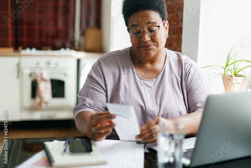 Portrait of puzzled upset serious overweight african american grandma in glasses reading paycheck or utility bill attentively sitting in her kitchen in front of laptop and smartphone photo
