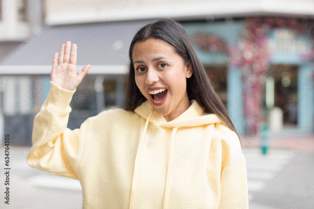 pretty hispanic woman smiling happily and cheerfully, waving hand, welcoming and greeting you, or saying goodbye