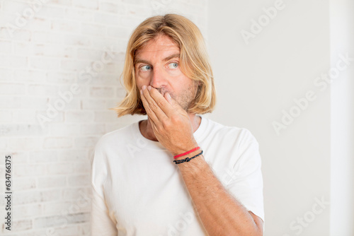 young blond adult man covering mouth with hands with a shocked, surprised expression, keeping a secret or saying oops