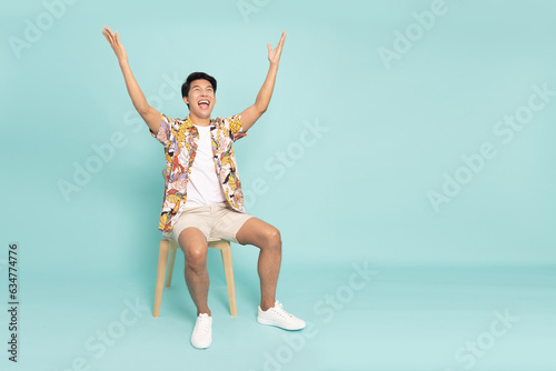 Attractive Asian man traveler sitting on white modern chair and hands up raised arms from happiness, Excited man blogger with winner success concept