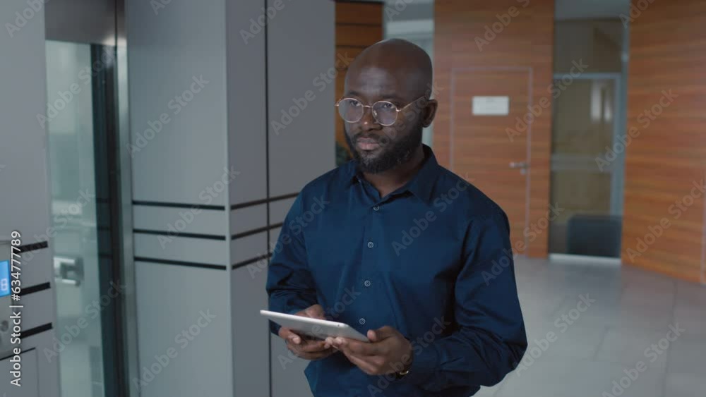 Medium shot of serious African American office worker using digital tablet while walking in hall of modern office Medium shot of serious African American office worker using digital tablet while walking in hall of modern office