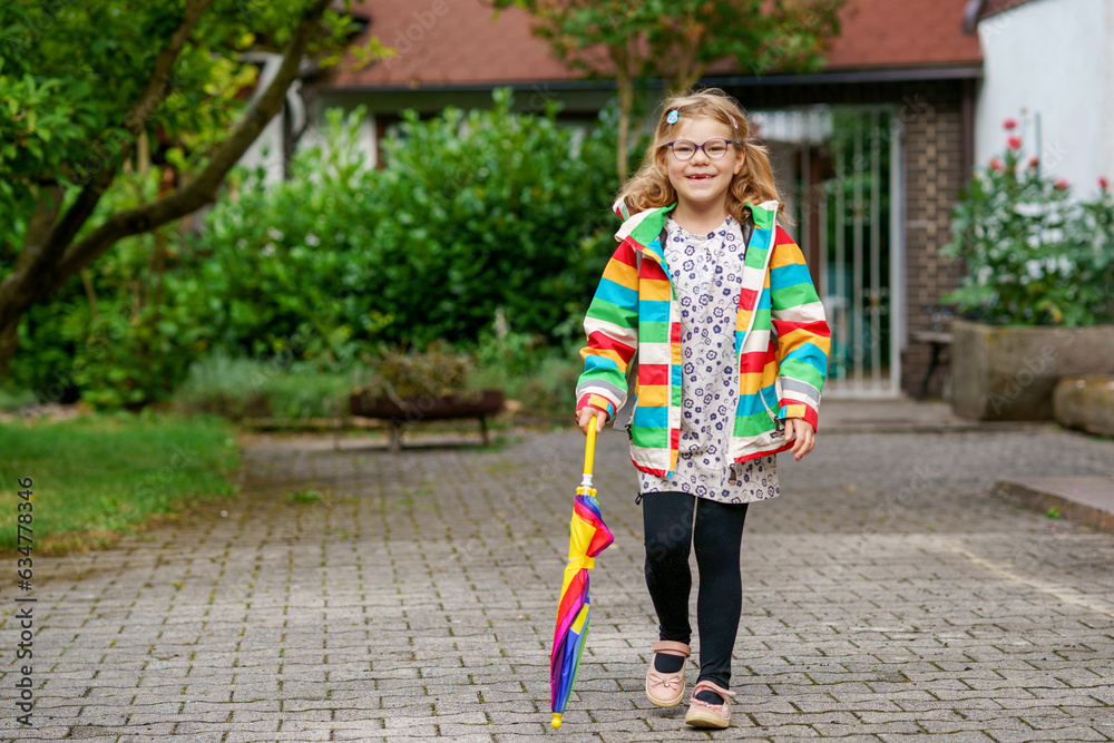 Little girl on way to elementary school or kindergarden. Preschool Child with colorful rainbow umbrella and waterproof jacket with school bag. Kid walking in autumn shower. Outdoor fun by any weather