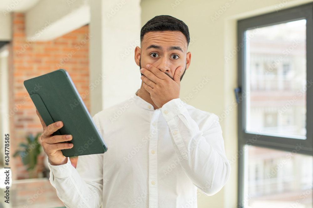 young adult man covering mouth with a hand and shocked or surprised expression. touchscreen pad concept young adult man covering mouth with a hand and shocked or surprised expression. touchscreen pad concept