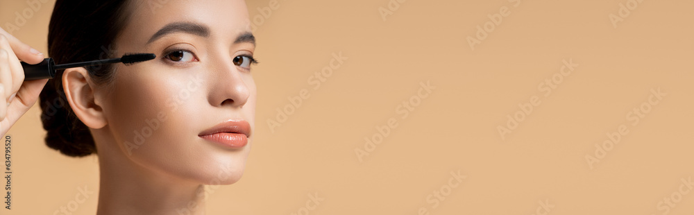 Pretty asian woman applying mascara and looking at camera isolated on beige, banner