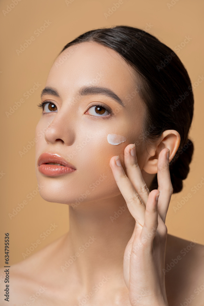 Young asian woman with naked shoulders applying face cream isolated on beige Young asian woman with naked shoulders applying face cream isolated on beige