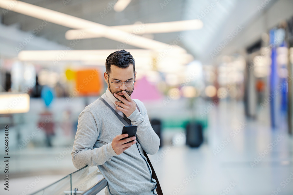 A young man is standing at the shopping mall and carefully reading messages on the phone. A young man is standing at the shopping mall and carefully reading messages on the phone.