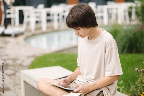 Teenage boy playing games on digital tablet and sitting on a bench under a house in a residential area