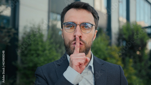 City portrait Caucasian business man businessman outdoors showing silence hush lips gesture keep secret secrecy confidential information stop talk silent quiet put finger to mouth mysterious hand sign