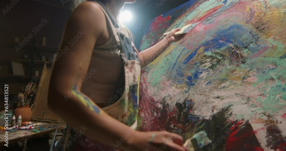 Millennial girl artist paints picture in loft studio using spatula to apply paint to canvas. Abstraction cinematic. Talented female artist energetically using spatula creates masterpiece of painting. 