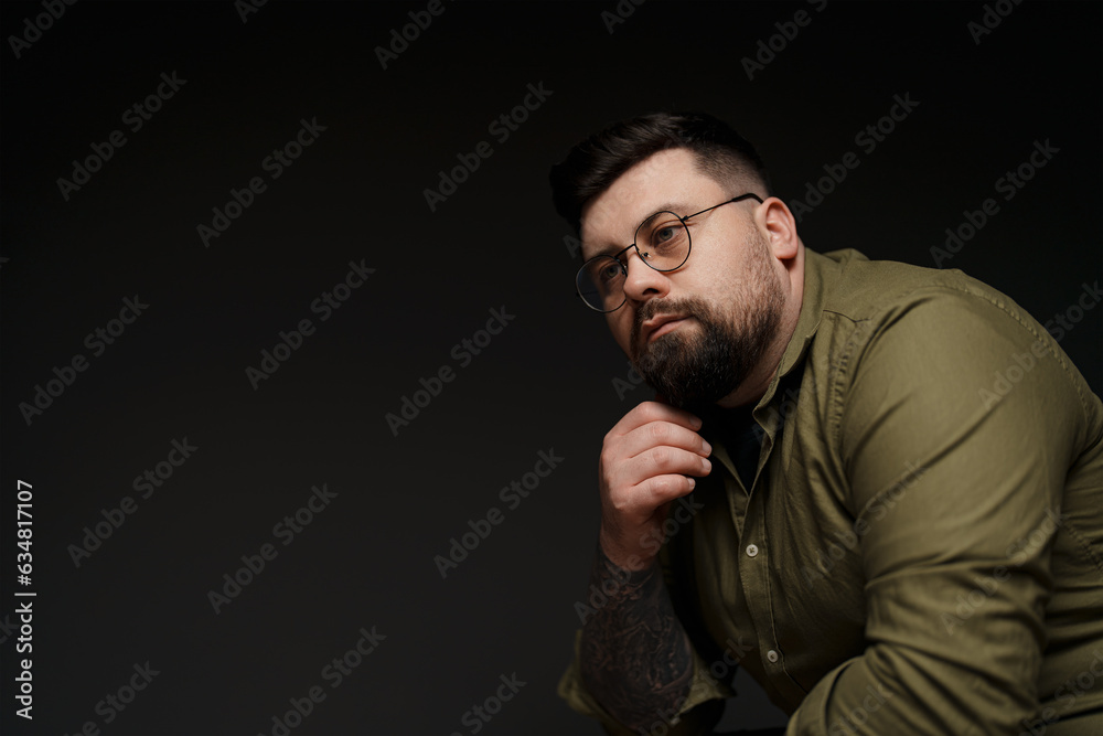 A portrait of a thoughtful bearded man in glasses wearing a green shirt. He is resting his chin on his hand and looking away from the camera. The background is dark gray