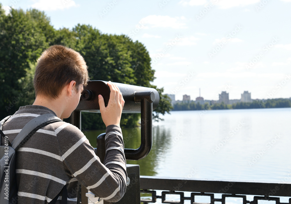 A teenager with a backpack looks in binoculars on river