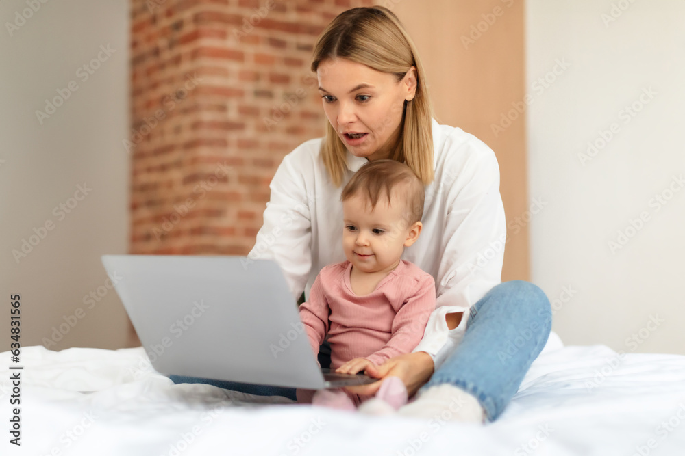 Baby and gadgets. Mommy and her little daughter using laptop, browsing internet for online fun, sitting on bed at home Baby and gadgets. Mommy and her little daughter using laptop, browsing internet for online fun, sitting on bed at home
