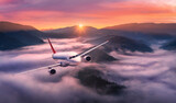 Airplane is flying above mountain peak in pink low clouds at sunrise. Landscape with passenger airplane, red sky, hills in fog. Aircraft is taking off. Business travel. Commercial plane. Aerial view