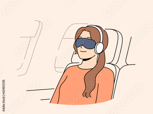 Woman passenger of flight with blindfold and earphones sleeps in airplane chair sitting outside porthole. Young girl first class passenger flies on business trip on airplane and smiles