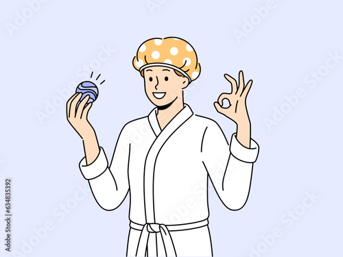 Man with bath bomb is dressed in bathrobe and bandage to protect hair from moisture. Young guy shows OK gesture recommending use of aromatic bath bomb while performing hygiene procedures.