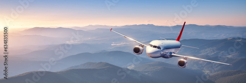 Plane is flying in colorful sky at sunset. Landscape with passenger airplane over mountains ranges and hills in fog, orange sky. Aircraft is landing. Business. Aerial view. Transport. Private Jetlane