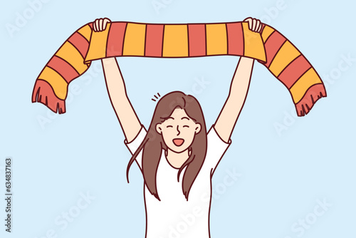 Woman soccer fan supports favorite sports team and shouts with joy raising scarf over head. Girl fan with clothes in colors of university team during tournament at football stadium.