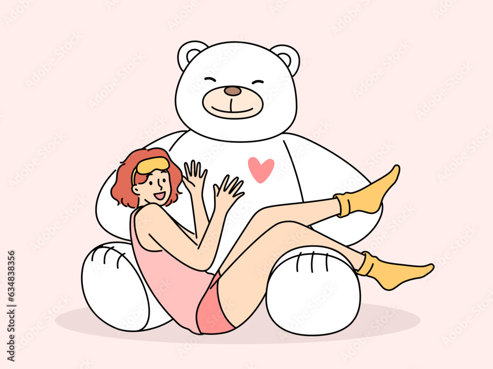 Giant teddy bear near sitting teenage girl in pajamas and sleep mask. Funny schoolgirl teen age looks at screen lying on feet of toy bear rejoicing at cute gift from parents or boyfriend - obrazy, fototapety, plakaty 