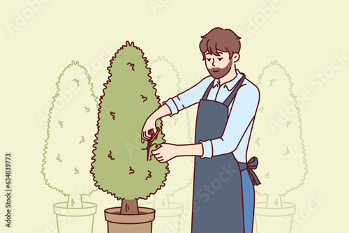 Man gardener cuts excess branches from trees growing in pots and taking care of plants to decorate landscape. Bearded guy gardener in work coat makes career as employee of greenhouse or conservatory