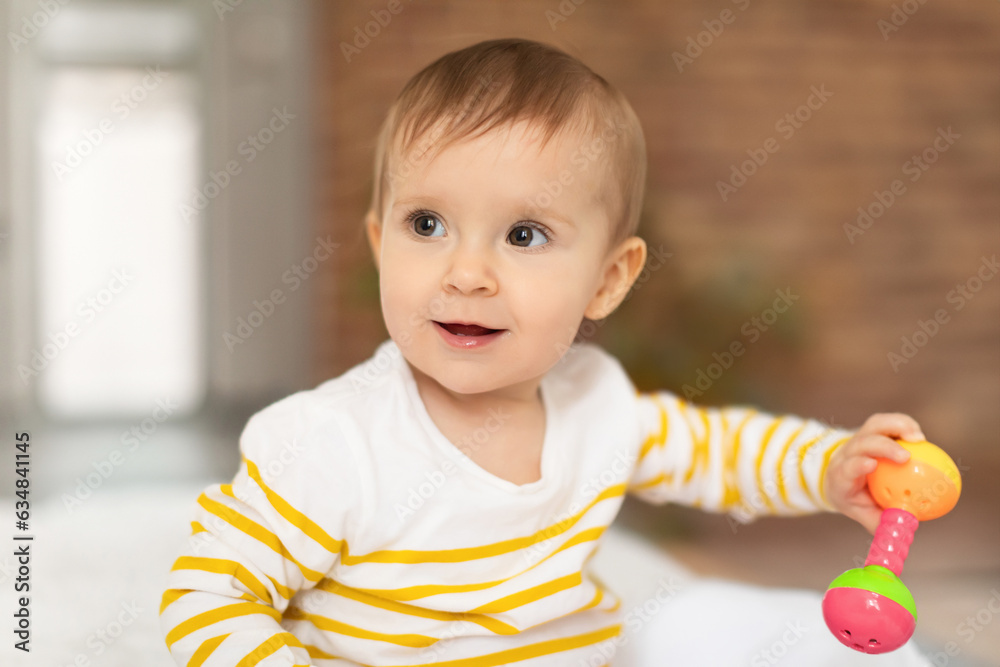 Infant baby girl playing with rattle toy while sitting on bed at home, looking aside and smiling Infant baby girl playing with rattle toy while sitting on bed at home, looking aside and smiling