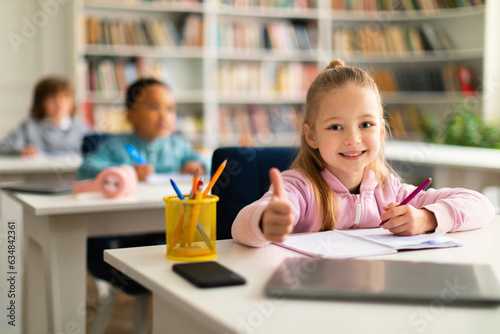 Smart girl doing thumbs up gesture while studying and sitting at desk in classroom  ready to study for excellence