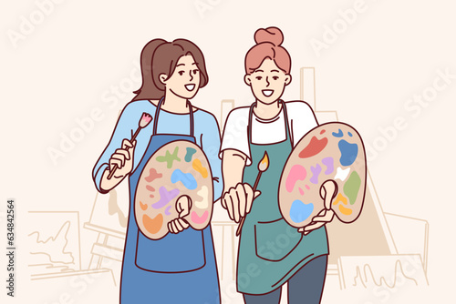 Two women artists hold brushes and palettes standing in workshop with easels to create artworks. Friendly girls creating artworks smiling looking at screen enjoying favorite hobby.