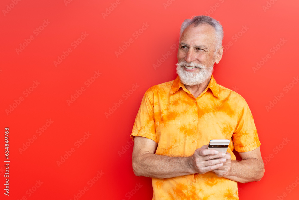 Handsome smiling senior man holding mobile phone shopping online on the street isolated on red background, copy space. Pensive bearded hipster using smartphone, communication online. Mobile banking 