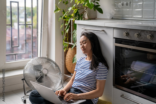 Relaxed Asian woman typing on laptop cooling by floor electric fan sits lotus position on floor in kitchen. Carefree Chinese girl blogger inspiredly writes article online magazine, work at home photo