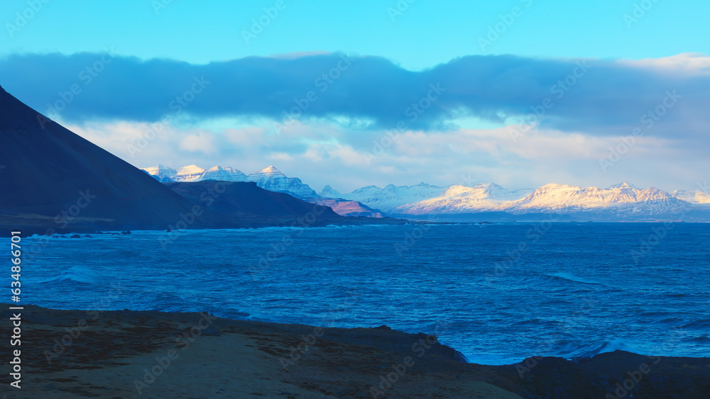 Majestic landscape of black sand beach in iceland, panoramic view of snowy mountains and freezing cold waters. Beautiful icelandic scenery on coastline shore with ocean waves. Handheld shot.