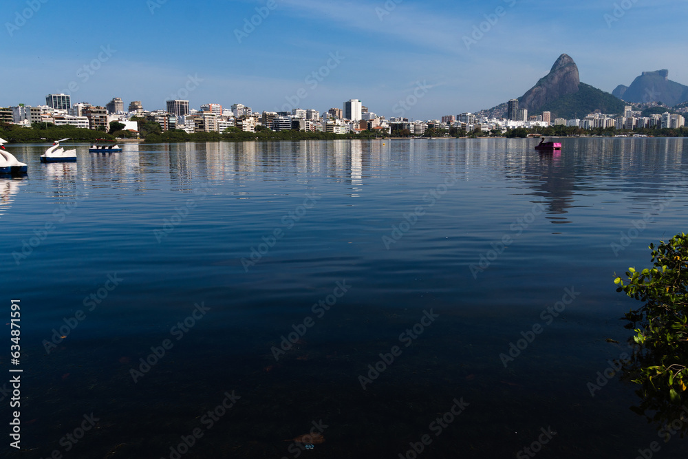 View of Rodrigo de Freitas Lagoon, south zone of Rio de Janeiro, Brazil. In the background, Dois Irmãos hill and Pedra da Gávea. Sunny day. Buildings around. Pond water used for sports