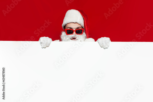 santa claus in hat and festive glasses shows empty banner for text on red background, man in santa costume