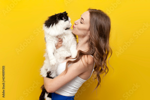 young cute girl kisses black and white cat on yellow isolated background and smiles, woman with pet photo