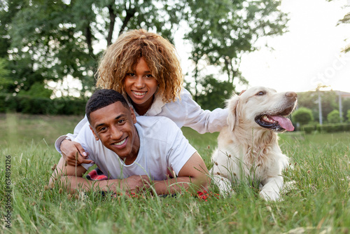 african american happy couple lie together with dog in park in summer, man and woman hug and love retriever