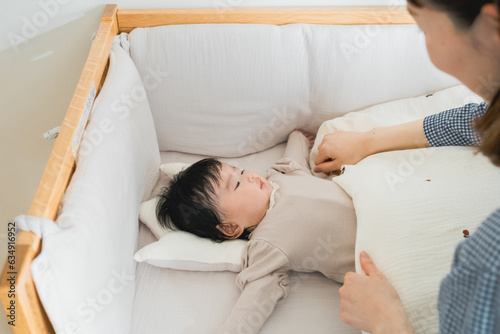 春の日中、ベビーベッドに横になる赤ちゃんの布団をかけ直す日本人の30代の女性