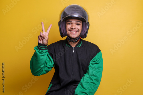 Asian online taxi driver wearing green jacket and helmet showing peace sign or two fingers over yellow background