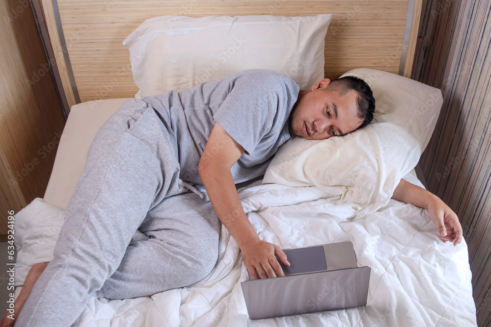 Sleepy lazy man working on laptop while sleeping on a bed Sleepy lazy man working on laptop while sleeping on a bed