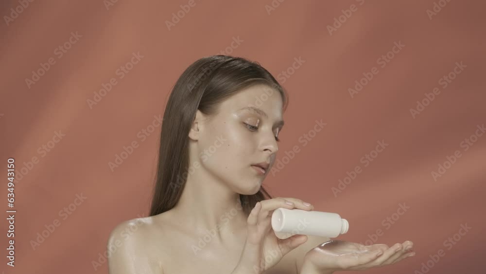 A woman pours lotion from a bottle into her palm and applies it to her bare shoulder. Portrait of a satisfied seminude woman in the studio on a red background. Slow motion. HDR BT2020 HLG Material.