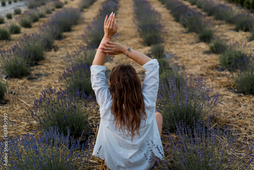 Fototapeta Naklejka Na Ścianę i Meble -  Rear view of a young woman in a casual shirt sitting and looking at lavender. The concept of relaxation and unity with nature