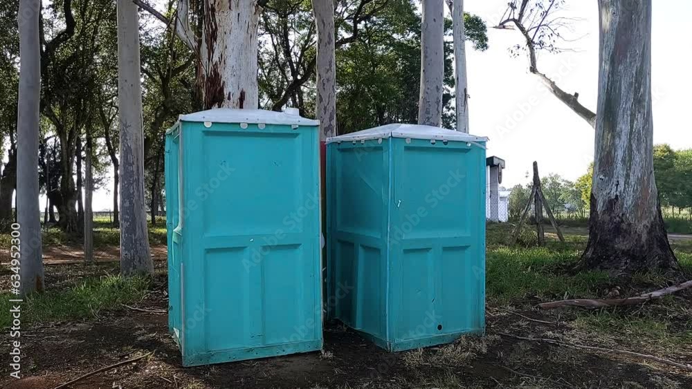 dolly forward of two aqua-green blue portable bathrooms in family park with trees dolly forward of two aqua-green blue portable bathrooms in family park with trees