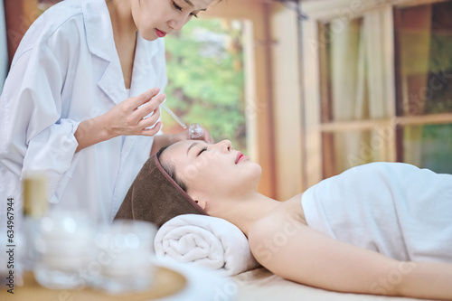 아시아 한국의 젊은 한국 여성 모델이 스파 또는 피부관리전문점 또는 피부과 병원에서 침대에 누워서 앰플 스포이드를 들고 있는 전문피부 관리사에게 피부 관리를 받고 있다.