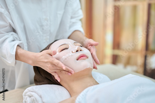 아시아 한국의 젊은 여성이 스파 또는 피부관리전문점 또는 피부과 병원에서 침대에 누워 눈을 감고 흰색 마스크팩으로 전문관리사에게 피부관리를 받고 있다.