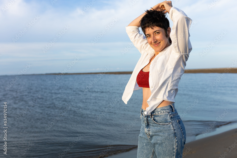 short-haired woman smiling on the beach, wearing white shirt, red top and denim, blue sky and calm sea.