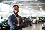 Portrait of confident young car dealer standing in showroom with arms crossed
