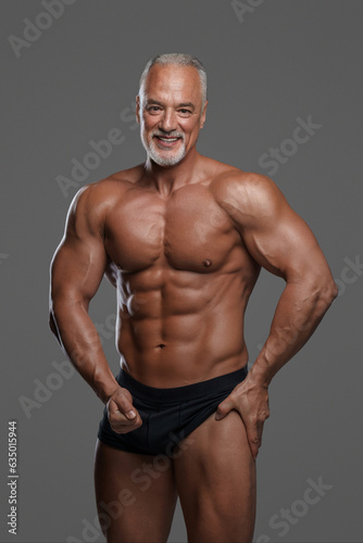 Charming mature muscular male model with stylish gray beard, wearing black underwear and posing with a big smile against a gray background © Fxquadro