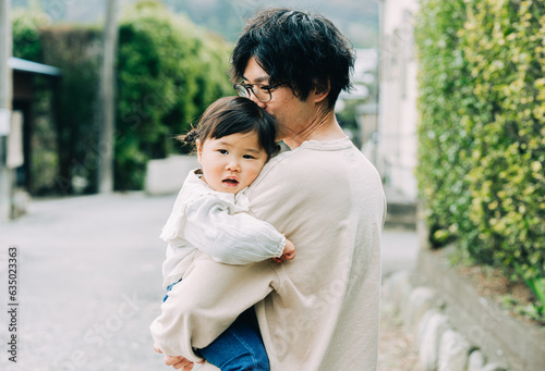 春の朝、屋外で小さい赤ちゃんを抱っこする眼鏡をかけた日本人の30代のお父さん
