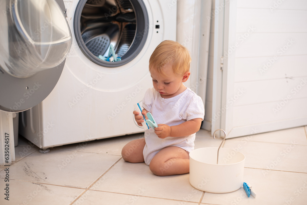 Little girl in white clothes sits on floor and play with clothespins near laundry machine. smile Baby put in side washing machine. First year of life, knowledge of world around. Little girl in white clothes sits on floor and play with clothespins near laundry machine. smile Baby put in side washing machine. First year of life, knowledge of world around.
