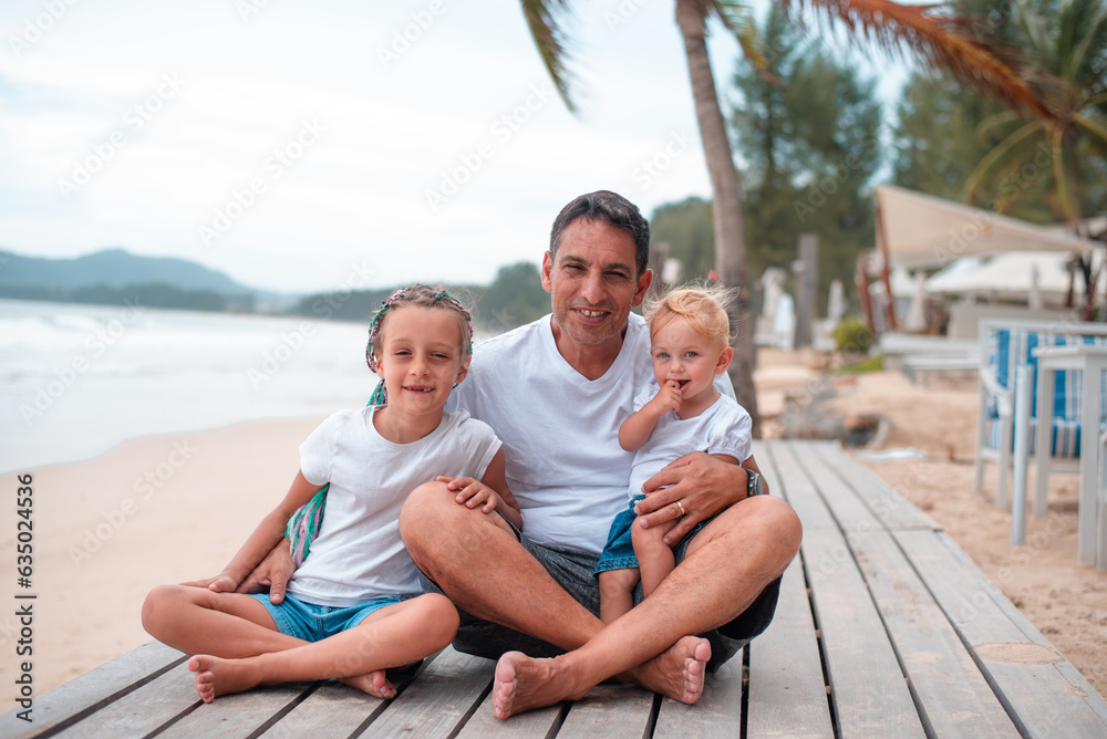 Summer vacation, happy family having fun. Happy smily two girls and father in middle sit on beach sea side in summer day. white clothes